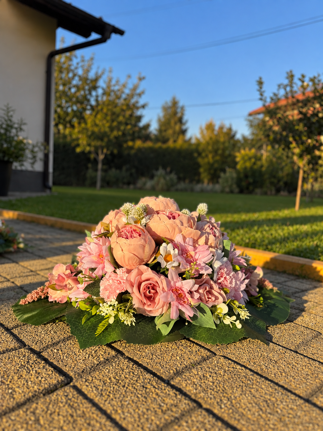 A large, vibrant artificial flower arrangement in shades of pink, peach, and green, displayed on a paved outdoor patio with a blurred garden background.